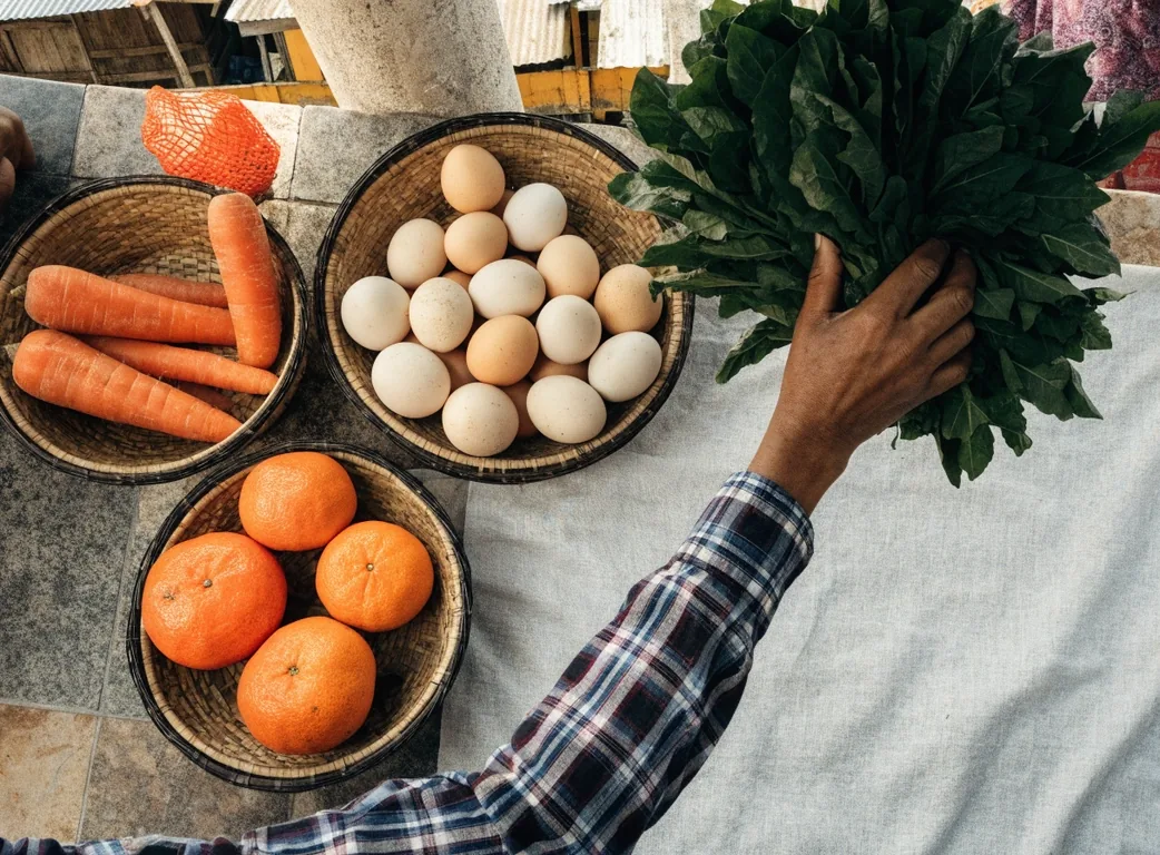 A flat overhead arrangement of market produce including greens, citrus, nuts and eggs on a bright ground
